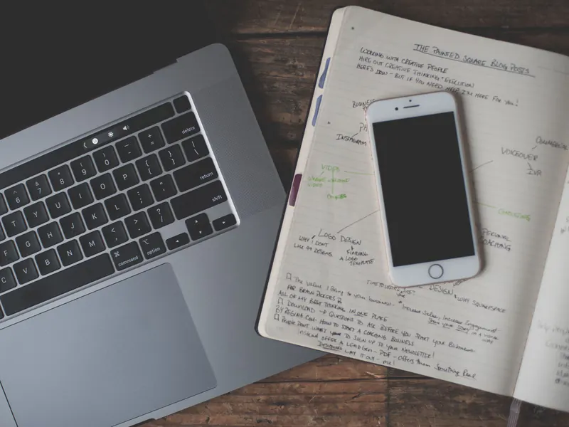 Overhead view of a laptop, smartphone and open notebook with handwritten blog and social media post ideas on a wooden desk