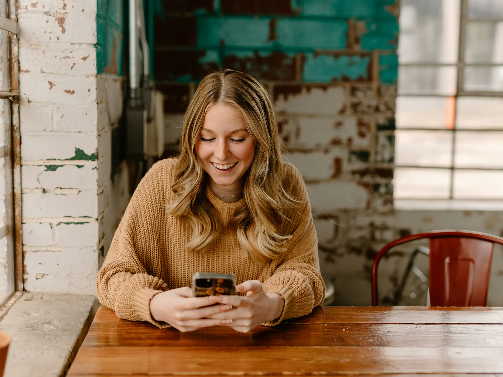 Smiling woman in a tan sweater scrolling her phone at a wooden table by the window of a brick-walled coffee shop