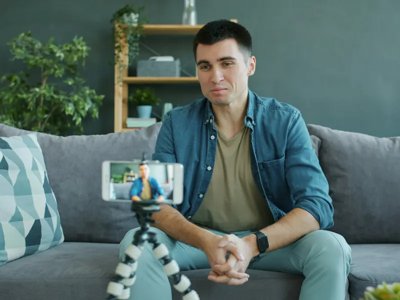 Man sitting on a sofa recording a video of himself on a smartphone mounted on a small tripod