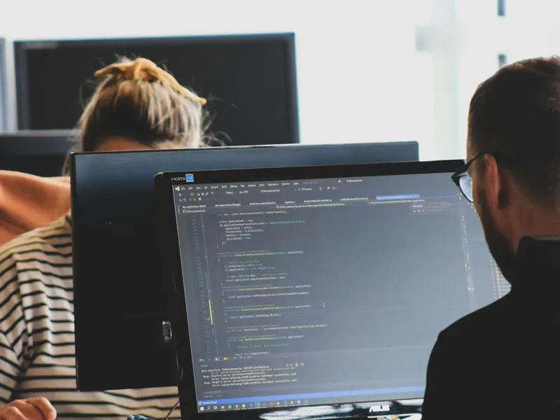 Two developers working side by side at monitors full of code in a bright office