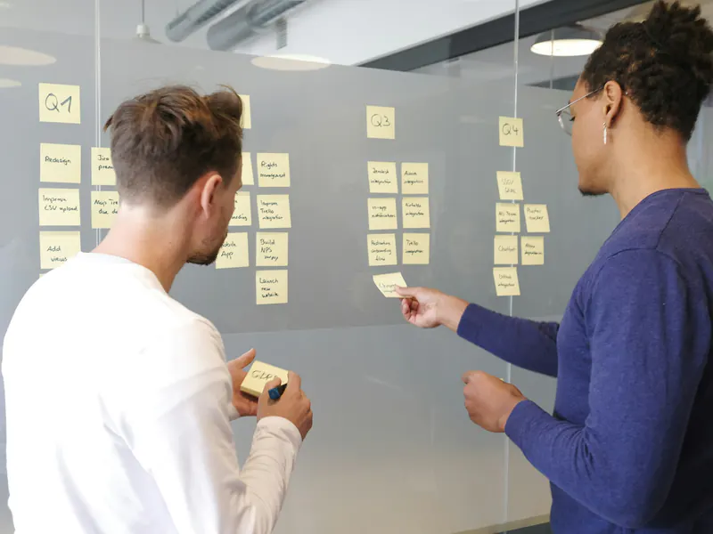 Two colleagues placing yellow sticky notes in quarterly columns on a glass wall while planning a project roadmap