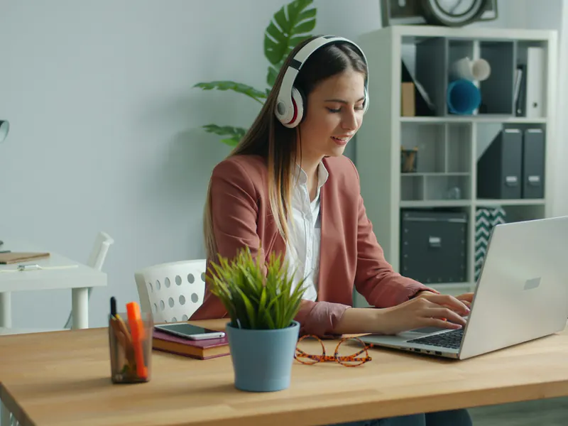 Woman wearing headphones typing on a laptop at a wooden desk in a bright home office with shelves and plants