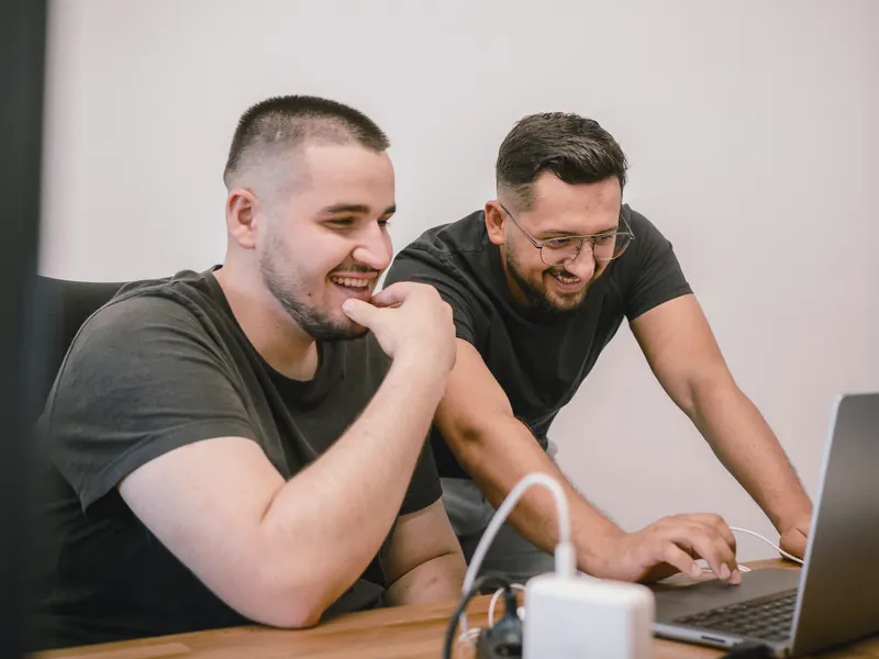 Two smiling colleagues in dark T-shirts leaning in to look at a laptop screen on a wooden desk