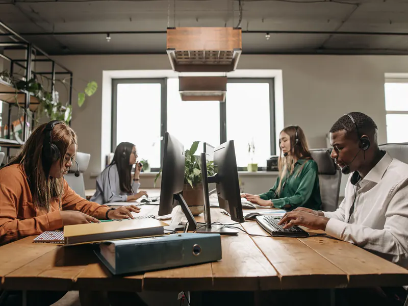 Four support staff wearing headsets work at computers along a shared wooden desk in a sunlit loft office