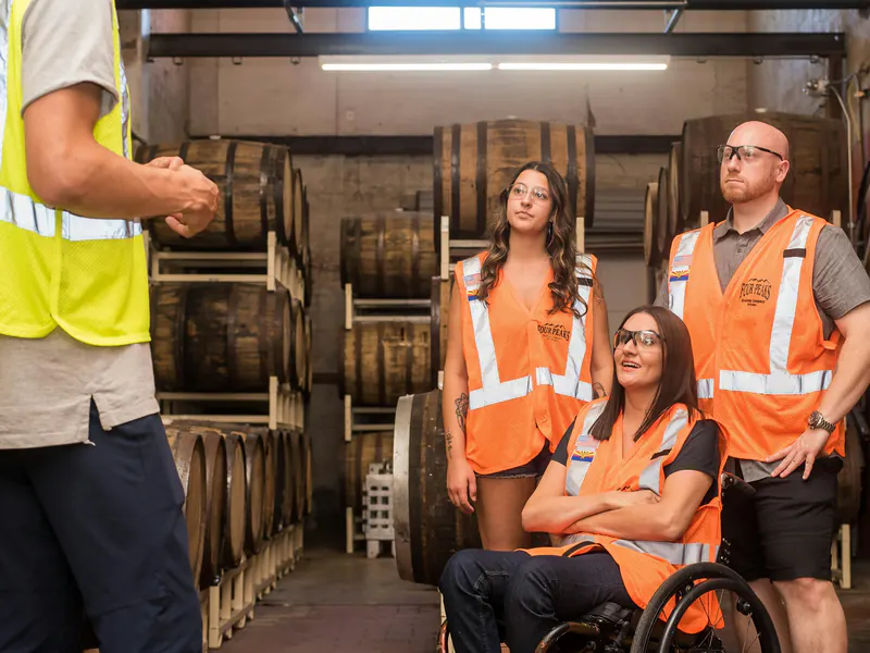 A supervisor in a yellow safety vest talks to three workers in orange vests, one in a wheelchair, in a barrel storage room.