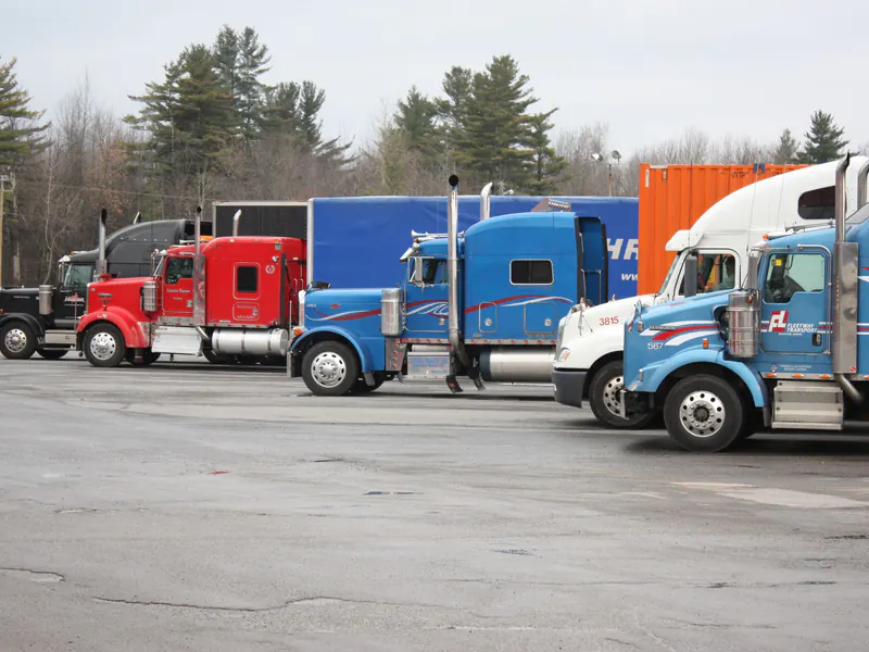 Row of parked semi tractors in red, blue, black and white lined up in an open paved lot with pine trees behind
