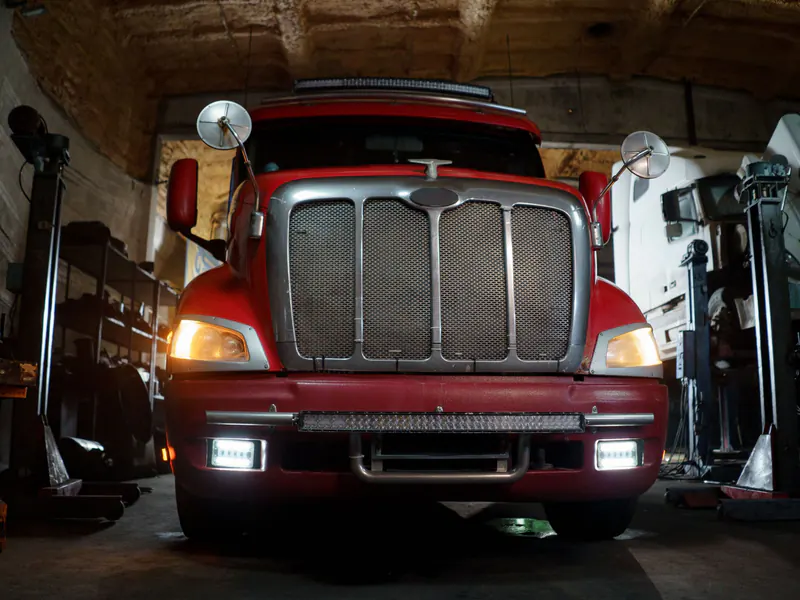 Front view of a red semi tractor with a chrome grille, bumper and lit headlights inside a dim repair garage