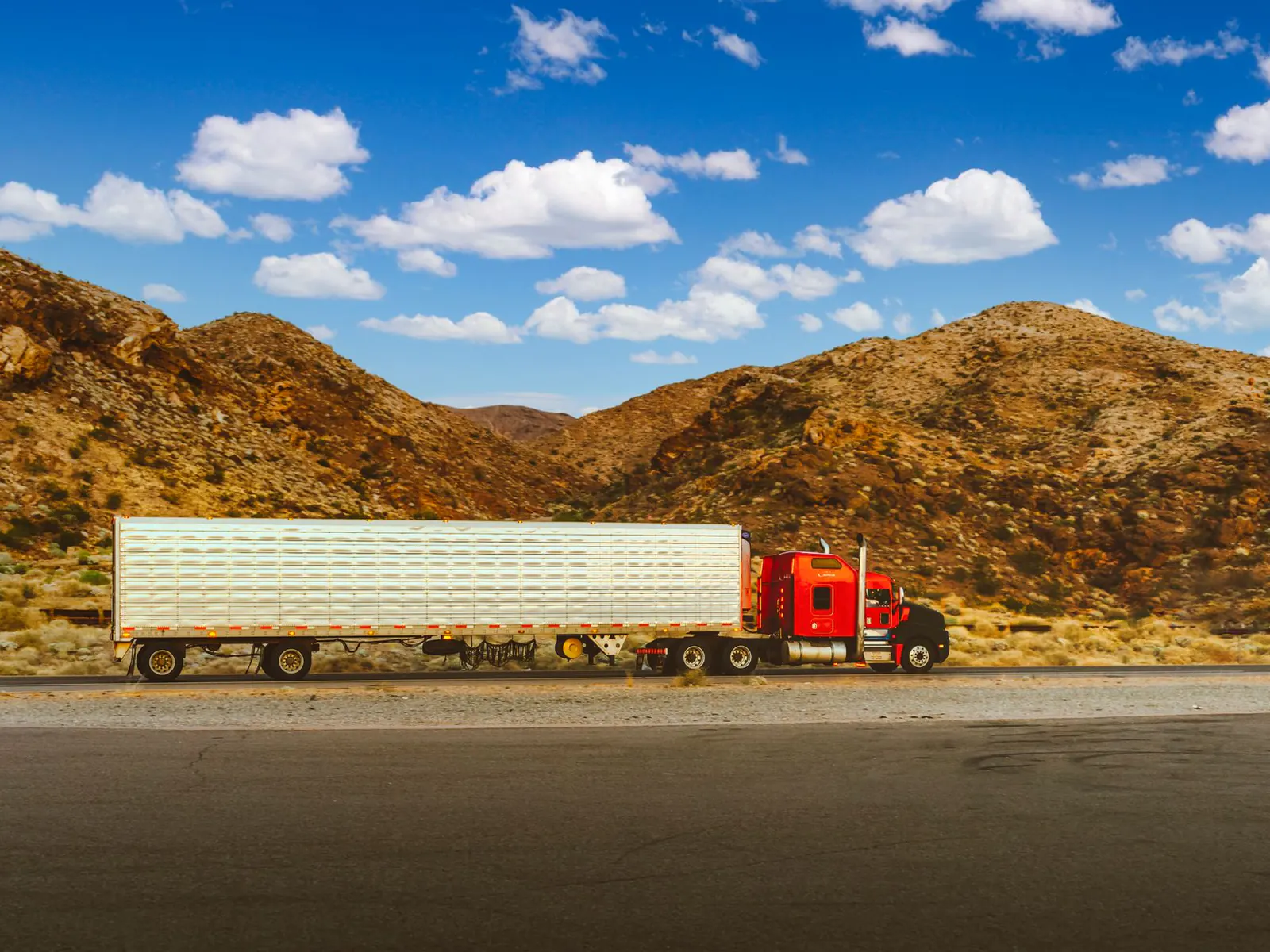 Red semi truck pulling a long white trailer along a desert highway below rocky hills and a blue sky in Nevada