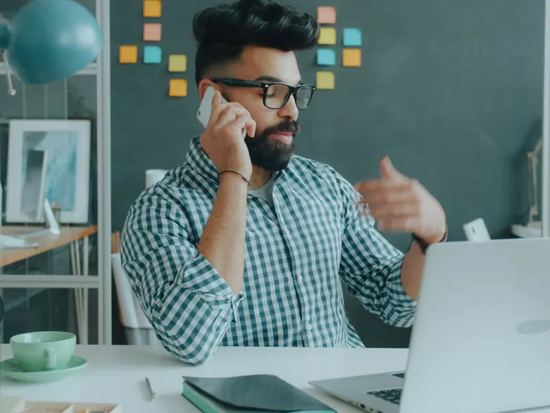 Man with glasses talking on his phone and gesturing while sitting at a desk with a laptop in a modern workspace