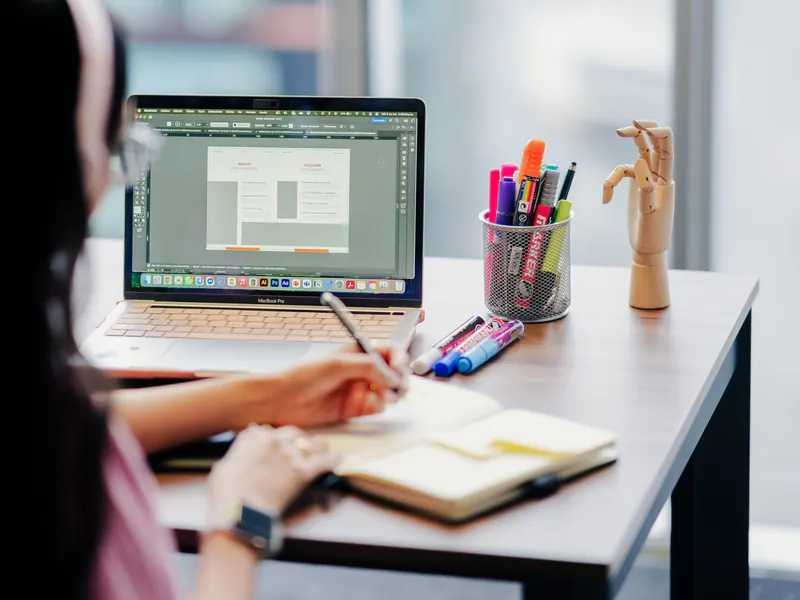Designer sketching layout ideas in a notebook beside an open laptop showing a page design, with a cup of markers on the desk