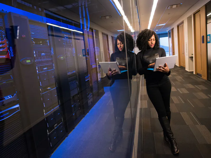 IT engineer holding a laptop beside glass-fronted server racks lit with blue light in a data center corridor