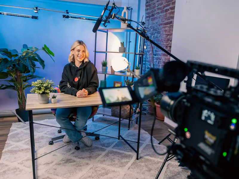 Woman sitting at a table in a blue-lit studio being filmed by a cinema camera with a boom microphone overhead